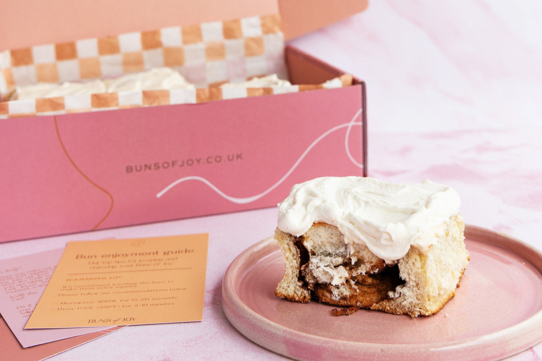 a single cinnamon bun on a pink plate next to a gift box with a personalised greetings card