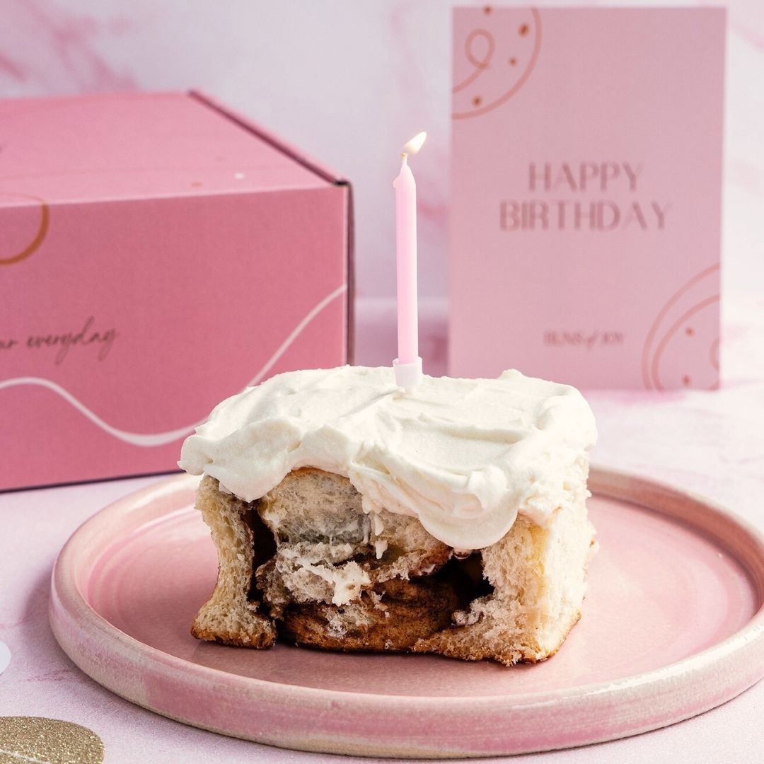 a single cinnamon bun on a plate with a candle in, with a happy birthday card behind