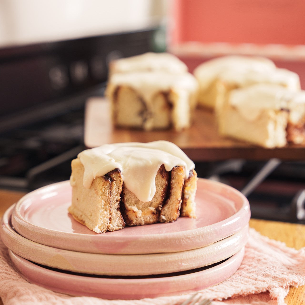 one cinnamon bun on a stack of pink plates with a group of 5 cinnamon buns on a board behind