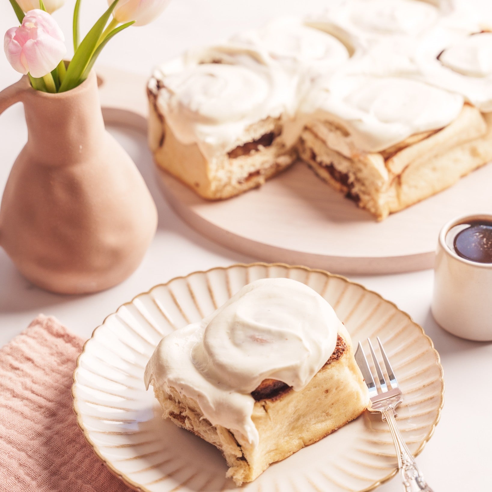 a cinnamon bun on a plate, next to a cup of coffee, with more cinnamon buns on a round board in the background